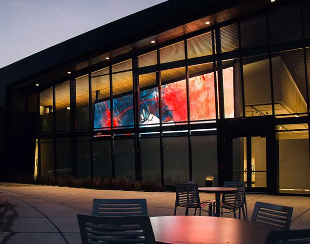 Large LED artwork display behind the glass façade of Mark Arts at night, with patio furniture in the foreground.