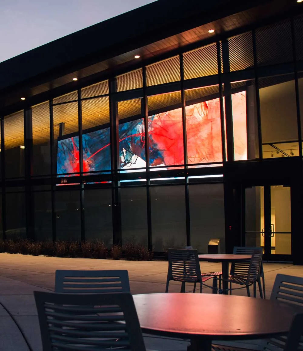 Modern building with large glass windows displaying colorful abstract art inside; outdoor patio area with empty metal chairs and tables in the foreground, illuminated by warm lights at dusk.