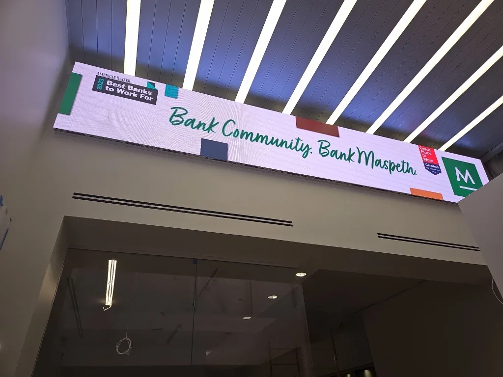 A wide indoor LED display at Maspeth Bank shows a community message beneath a ceiling with illuminated linear strips.