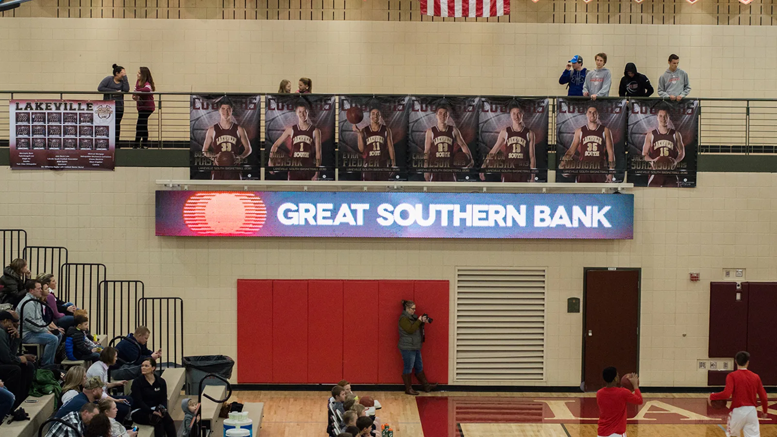Wide LED display showing Great Southern Bank branding on a gym wall below basketball player banners, with people on an upper balcony and a photographer below.