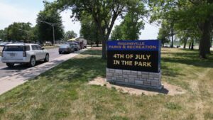 An LED electronic message center in a grassy park displays “Higginsville Parks & Recreation: 4th of July in the Park.” Cars and trees nearby.