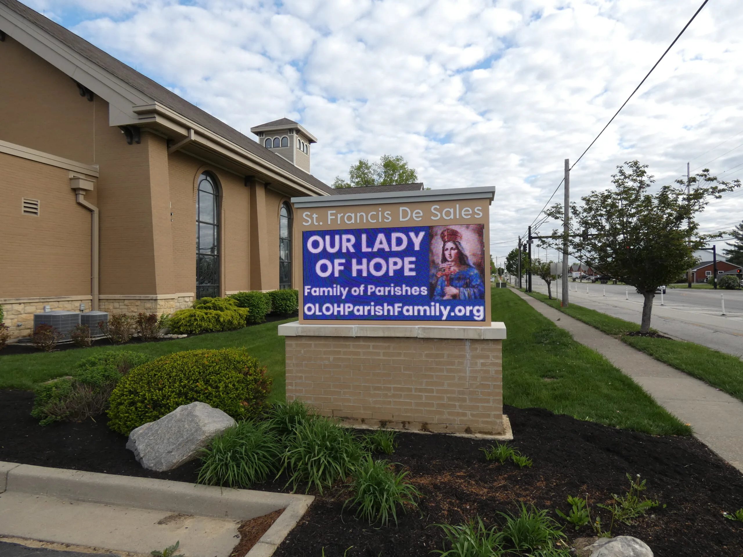Low monument sign beside the church building shows an Our Lady of Hope message on its LED display, with landscaping and a sidewalk nearby.