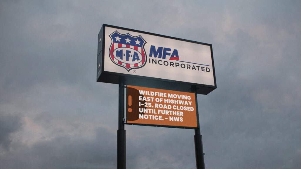 An LED electronic message center below the MFA Incorporated logo alerts: Wildfire by I-25, road closed, NWS update; cloudy sky backdrop.