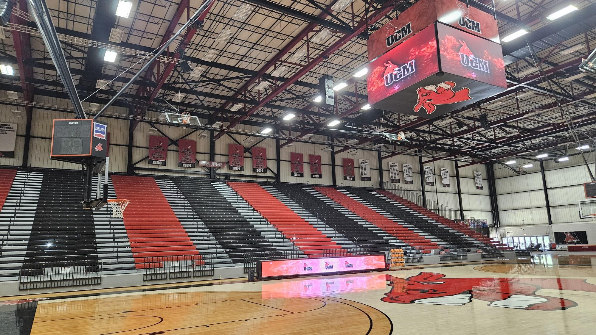 Spacious basketball gym with red, black, and white bleachers, banners, an LED electronic scoreboard showing UCM, and a red mule logo at center court.