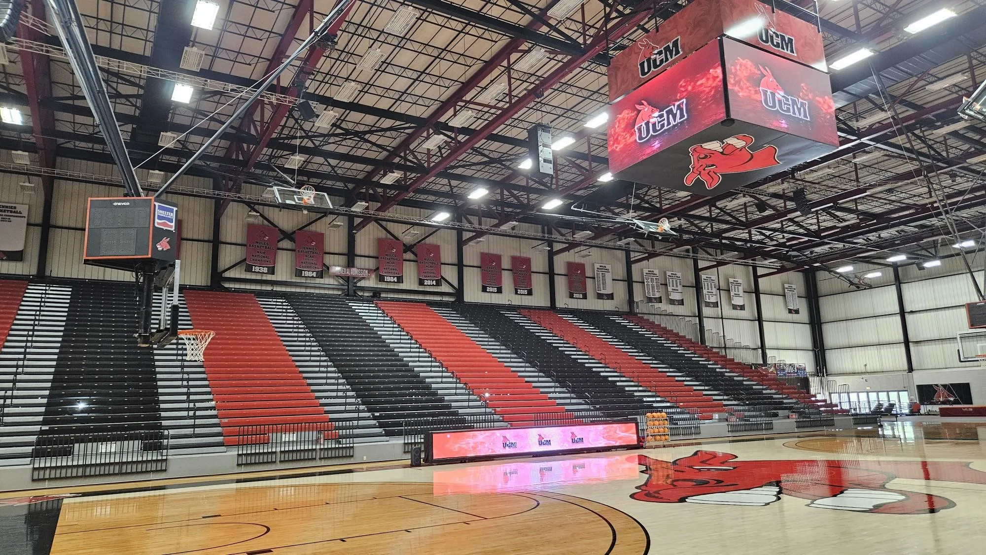 Spacious basketball gym with red, black, and white bleachers, banners, an LED electronic scoreboard showing UCM, and a red mule logo at center court.