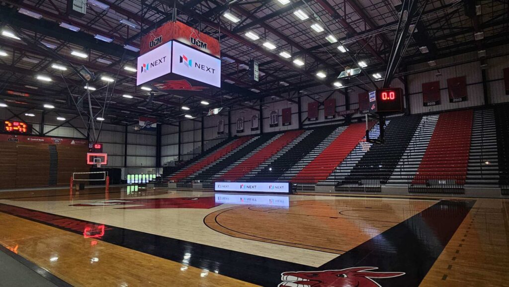 Empty basketball court with a solid-state LED scoreboard, aluminum-framed electronic message centers, and red, black, white bleacher seating.