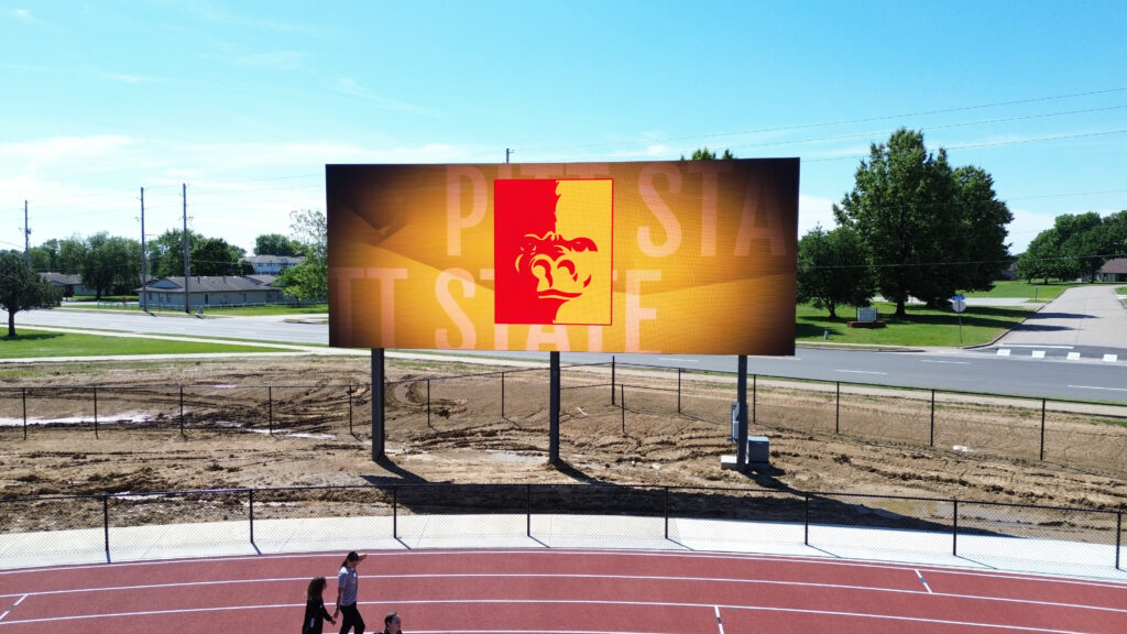 A high-brightness LED electronic message center shows red and yellow graphics with mascot face, above track, roadway, trees, and buildings.