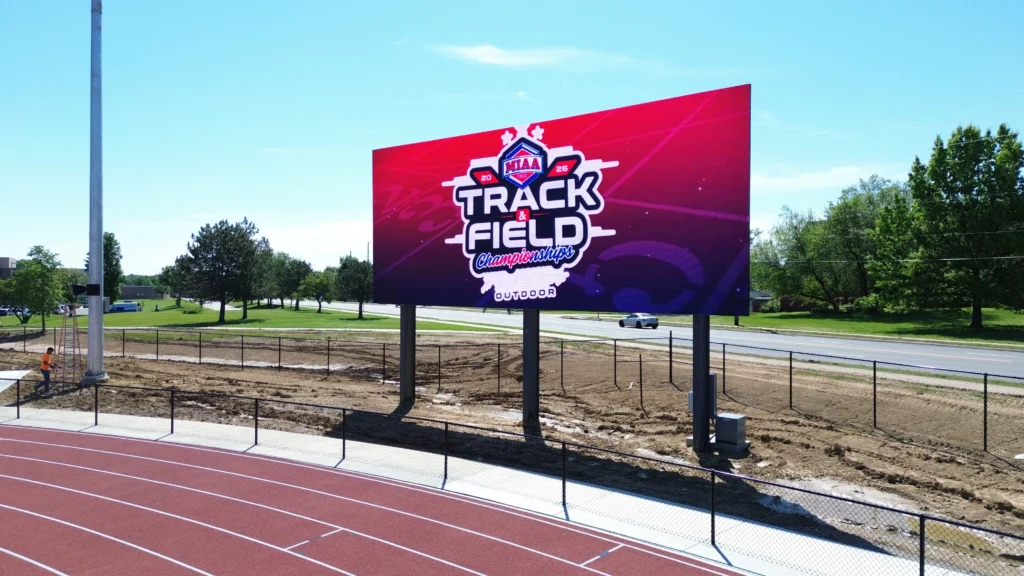 High-brightness LED electronic message center by a track displays “NAIA Track & Field Championships Outdoor,” with trees and road behind.