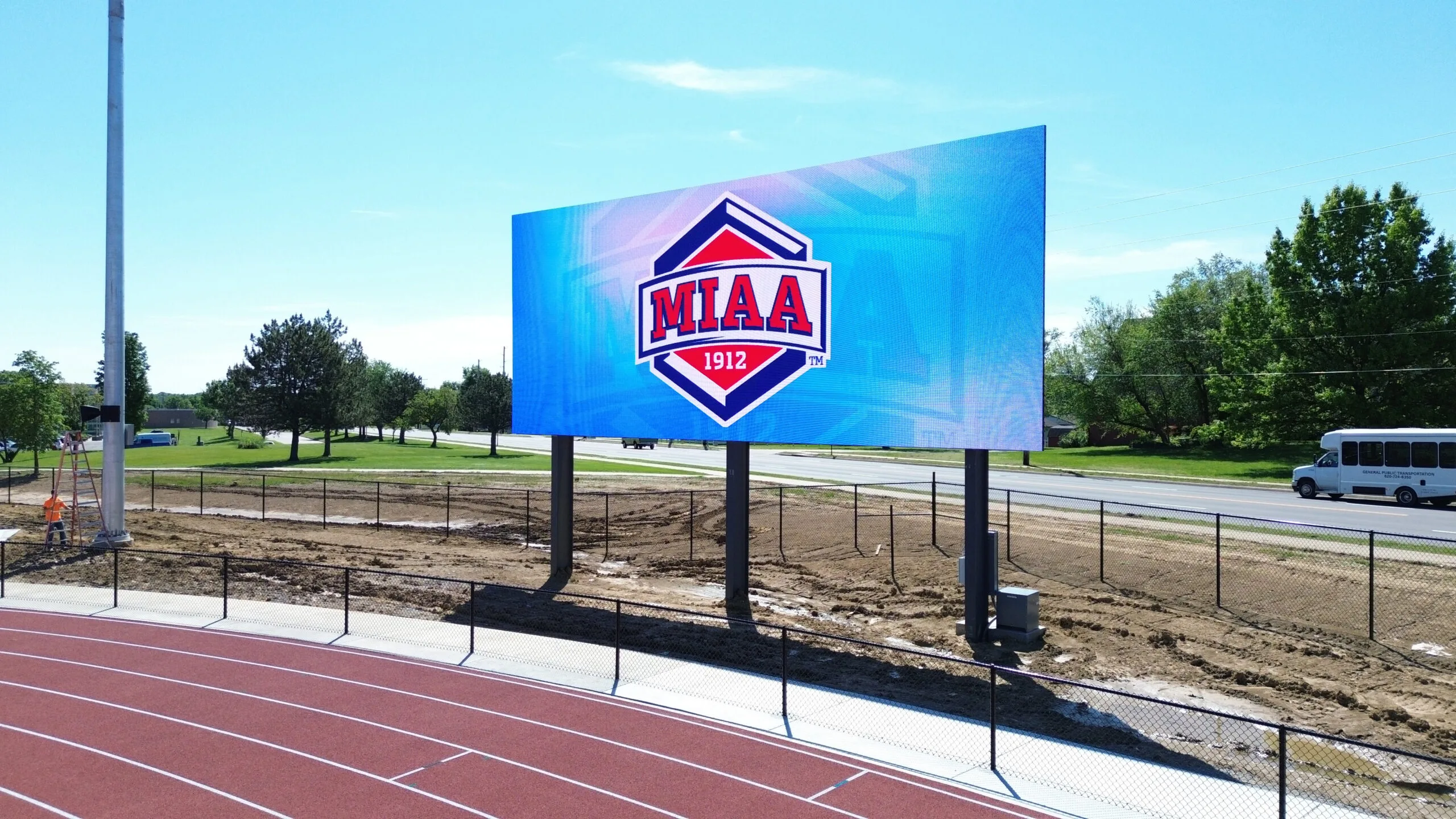 High-brightness LED electronic message center with MIAA 1912 logo by a running track; road, trees, and white bus in background.