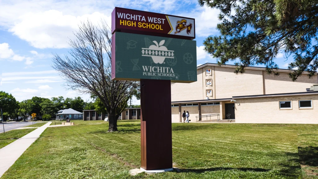An LED electronic message center for Wichita West High School on a grassy lawn, in front of a tan brick building with tree and sidewalk.