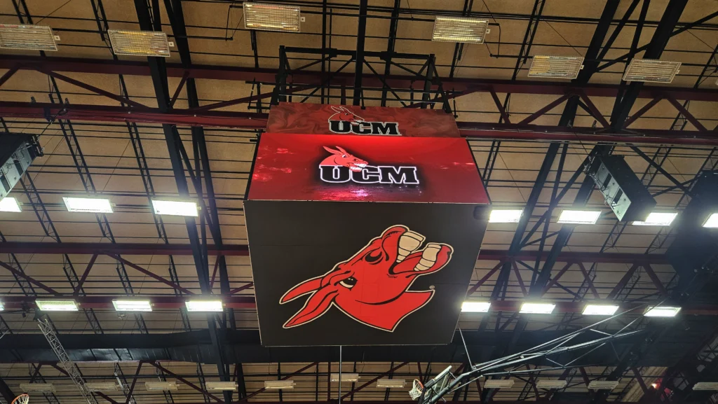 Ceiling-mounted LED sign in arena shows UCM mule head logo in red and black; high-brightness display, aluminum frame, beams above.