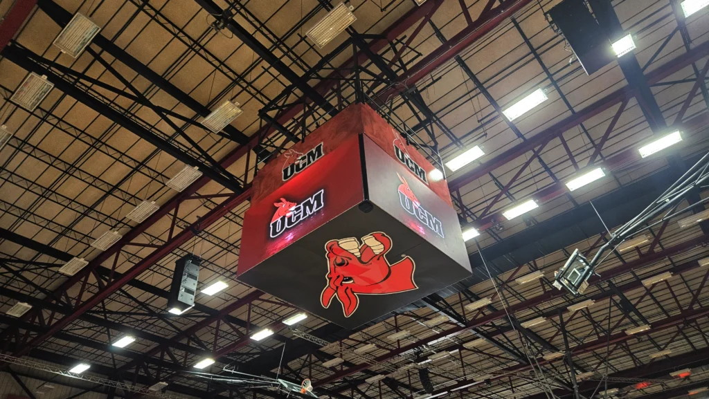 High-brightness LED electronic message center with UCAM logo and red bull mascot, suspended from arena ceiling with metal beams.