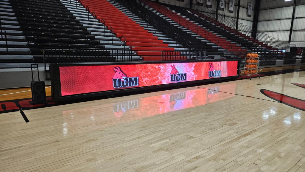 Basketball court with high-brightness LED sign showing UCM logo, in front of black and red bleachers; basketballs stacked at right.