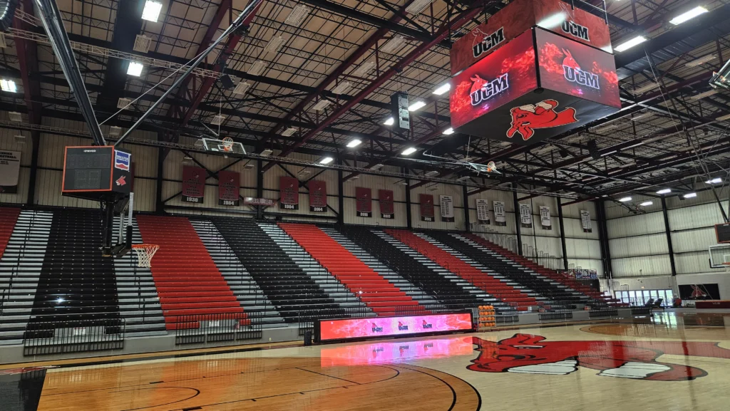 Empty basketball arena with black, red, and white seats, high-brightness LED scoreboard, banners, UCM logos, and red mule court mascot.