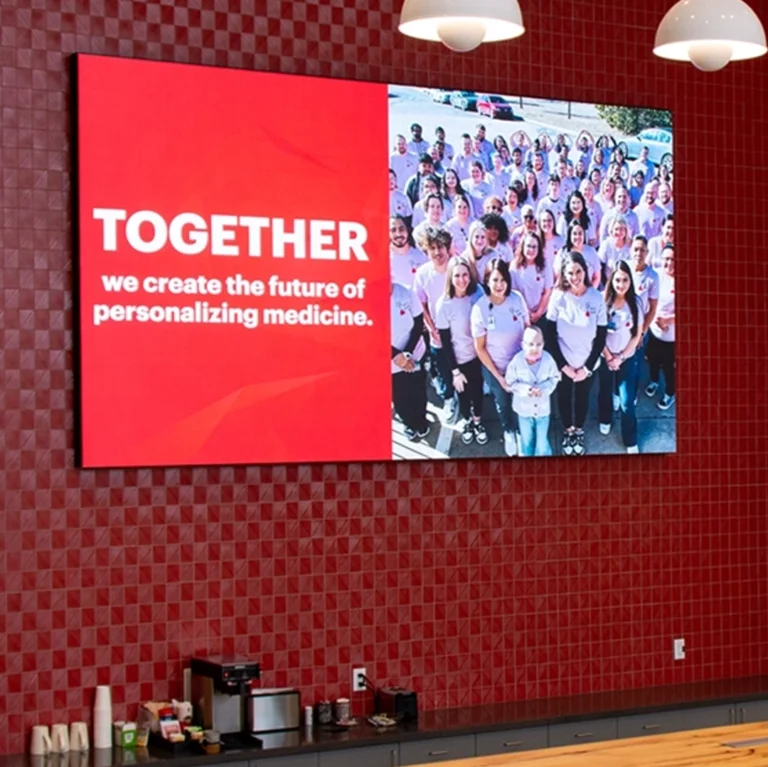 High-brightness LED sign shows group photo and "TOGETHER we create the future of personalizing medicine," on red-tiled wall above coffee station.