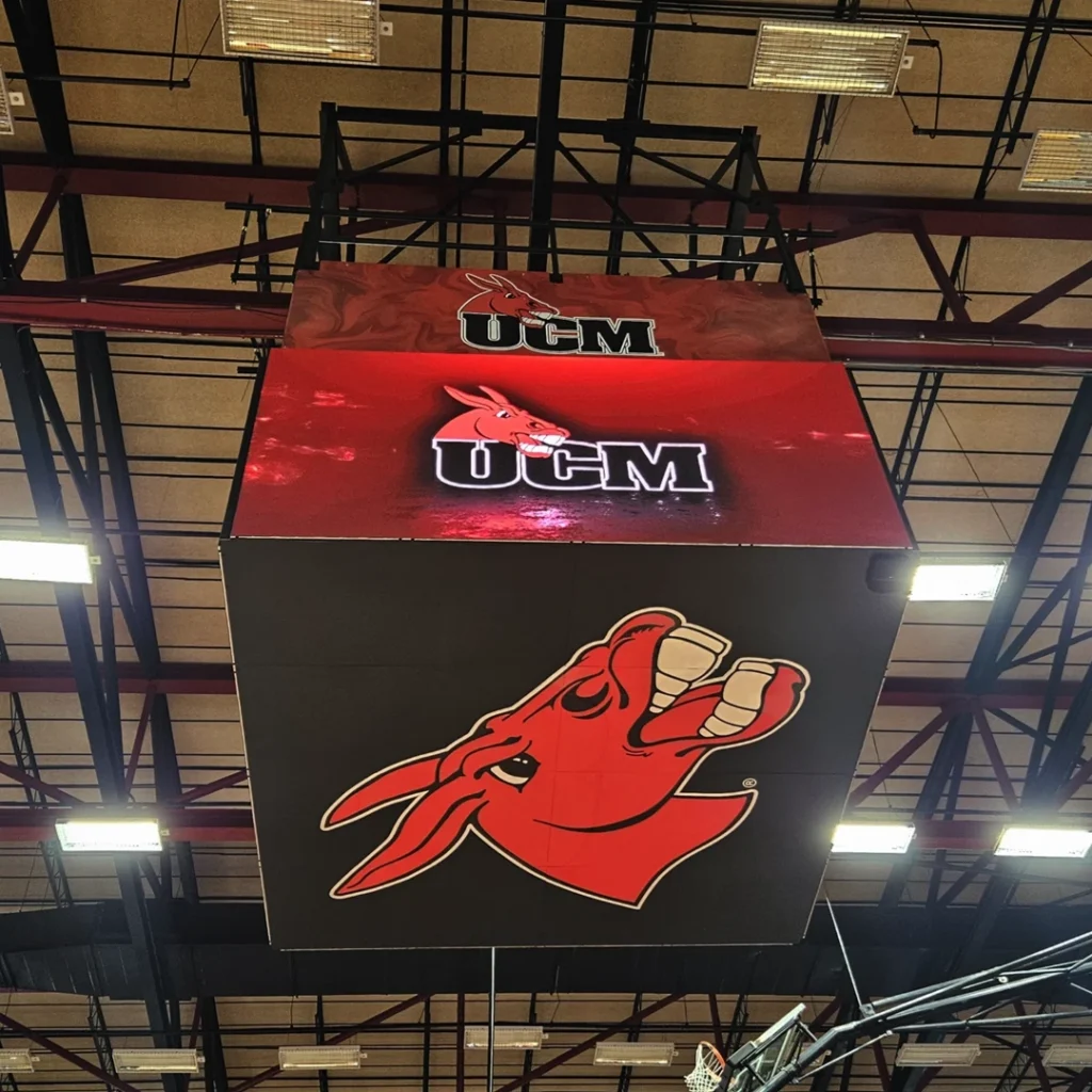 High-brightness LED message center with red mule mascot and bold “UCM” in white, suspended from a gym ceiling with metal beams.