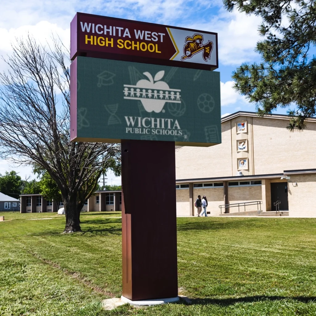 An LED electronic message center displays the Wichita Public Schools logo outside beige West High; two people walk nearby, trees visible.