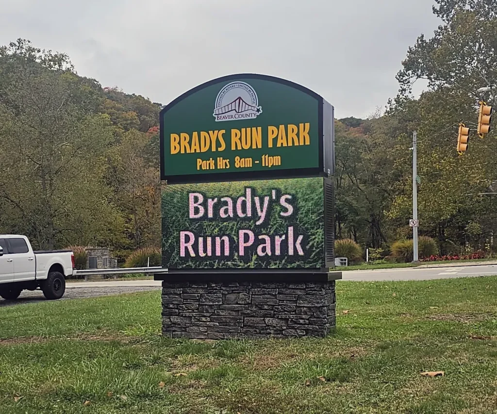 An LED electronic message center at Bradys Run Park displays park hours as 8am to 11pm. The solid-state, high-brightness sign is positioned on a grassy area with trees, a white truck, and a traffic light in the background.