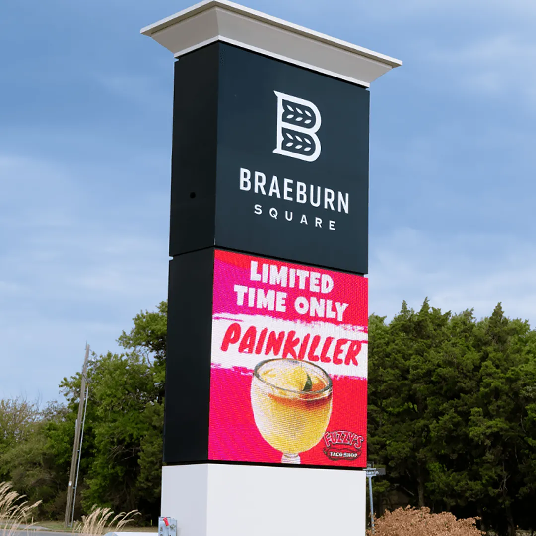 Braeburn Square features a tall commercial sign with an aluminum frame, topped by an LED electronic message center displaying a high-brightness digital ad for Fuzzy’s Taco Shop. The screen shows a cocktail image and the text “LIMITED TIME ONLY PAINKILLER.” Trees and clear blue sky are visible in the background.