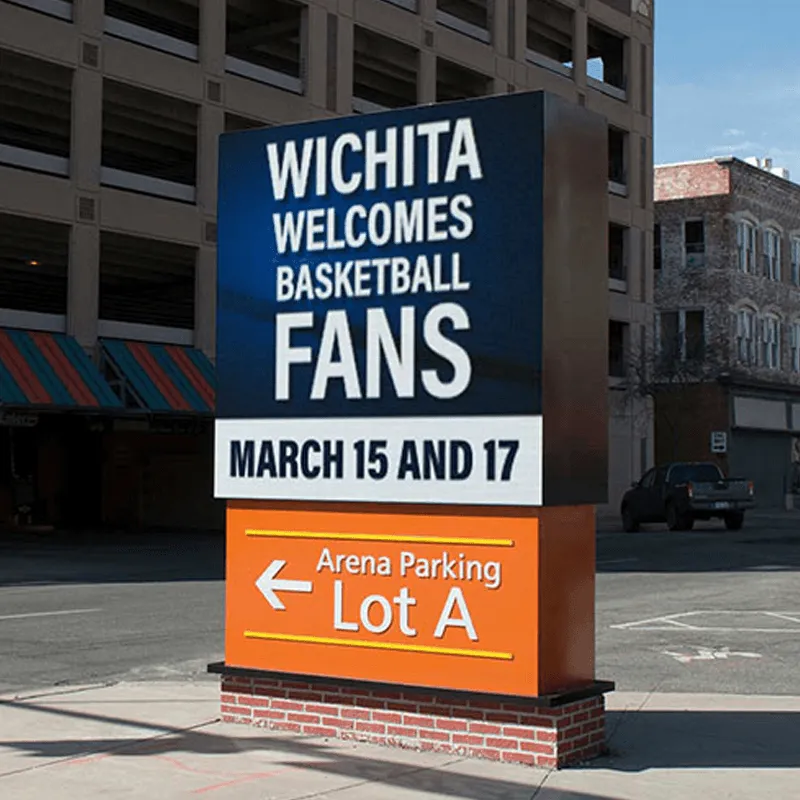 LED electronic message center displays: “Wichita welcomes basketball fans March 15 and 17.” Below, a left-pointing arrow and text indicate “Arena Parking Lot A.” The solid-state sign is positioned before a parking garage and adjacent buildings.