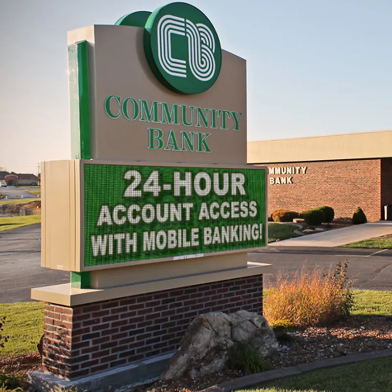 An LED electronic message center in front of a brick Community Bank building displays “24-HOUR ACCOUNT ACCESS WITH MOBILE BANKING!” in bright text. The sign is set amid grass and pavement.