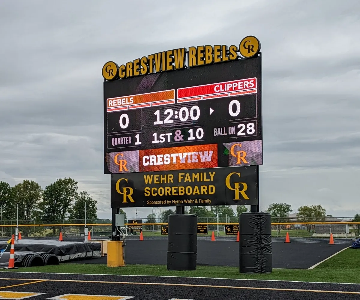 An LED electronic message center at Crestview Rebels field shows a 0-0 score for Rebels vs. Clippers, quarter 1, with 12:00 remaining. The scoreboard features high-brightness digits, the team logo, and “WEHR FAMILY SCOREBOARD” on the solid-state display cabinet below.