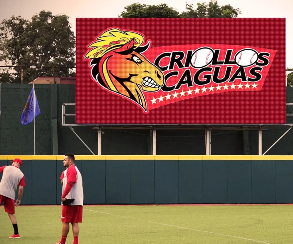 Two baseball players stand on a field in front of an LED scoreboard with a horse logo and “Criollos Caguas” text above star graphics. The electronic message center features high-brightness display elements. Trees and a blue flag are visible in the background.