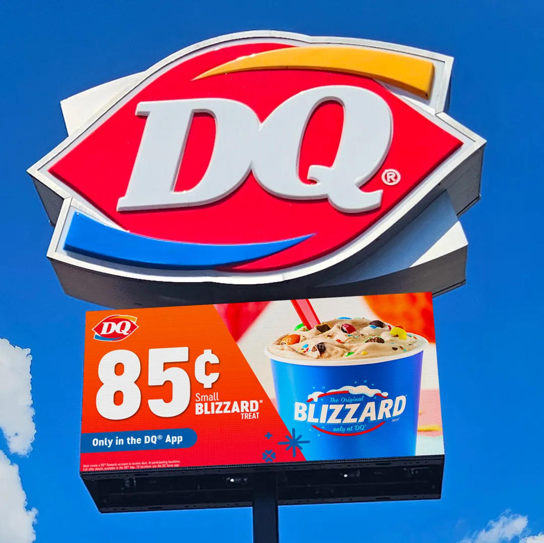 Large DQ sign mounted above an LED electronic message center displaying a high-brightness image of a small Blizzard treat in a blue cup, with the promotion “85 cents” and offer details, set against a partly cloudy blue sky.