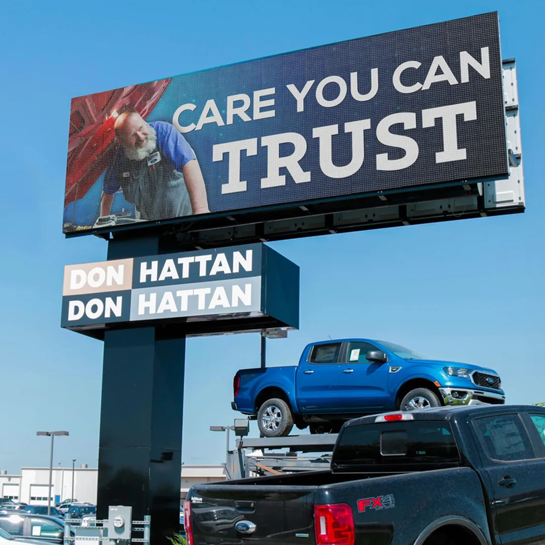 An LED billboard displays the message CARE YOU CAN TRUST alongside a mechanic graphic. The electronic message center shows DON HATTAN twice below. At the car dealership lot, a blue truck is positioned above a black truck.