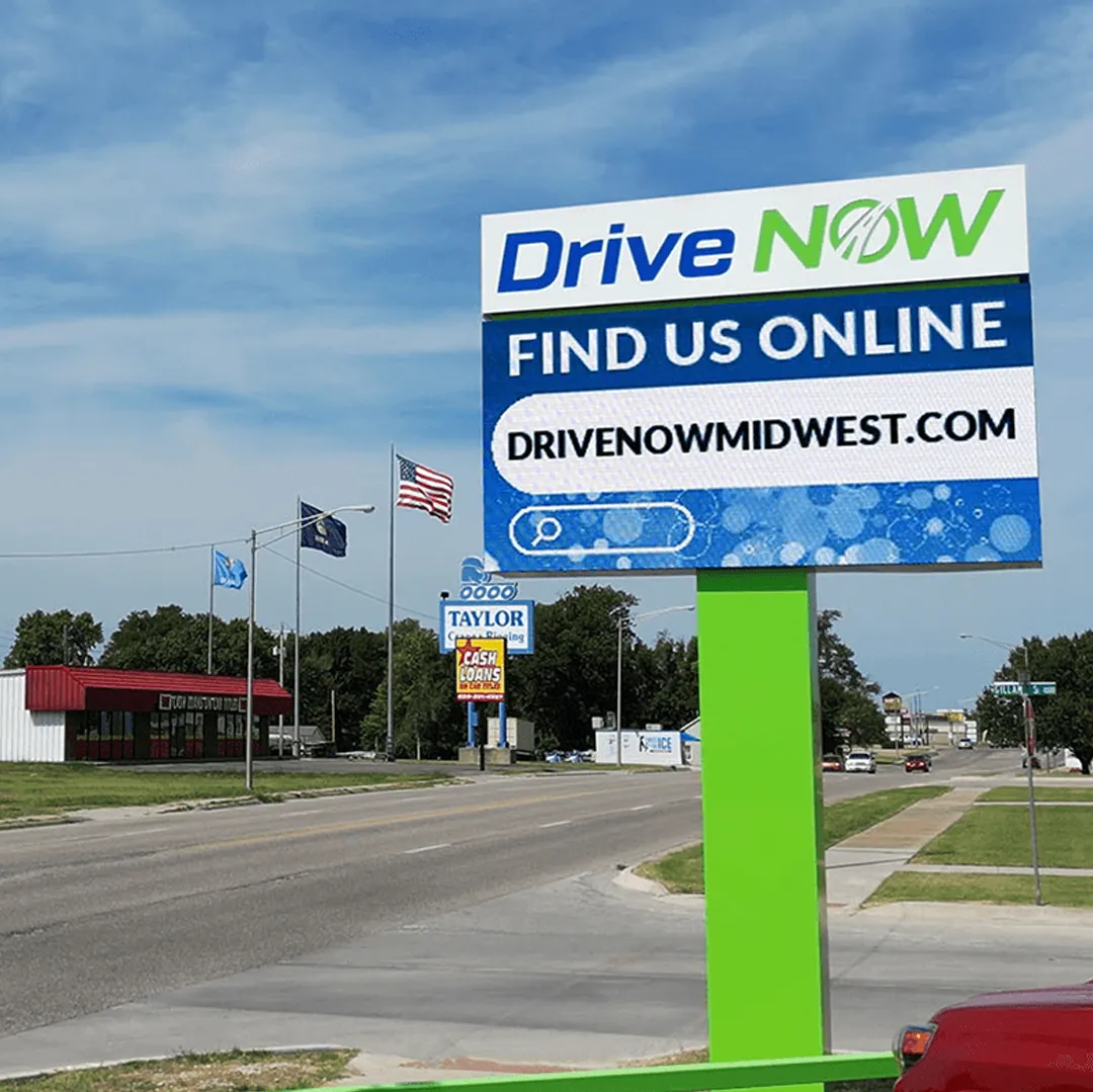 STAX an LED electronic message center displays "Drive Now. Find Us Online. DRIVENOWMIDWEST.COM" along a suburban street with visible businesses, vehicle traffic, and a partly cloudy sky.
