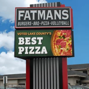 Restaurant signage displays “FATMANS Burgers • BBQ • Pizza • Volleyball” in bold letters above a high-brightness LED electronic message center. The digital sign shows an image of pizza with the message “Voted Lake County’s Best Pizza.” Blue sky and building appear in the background.