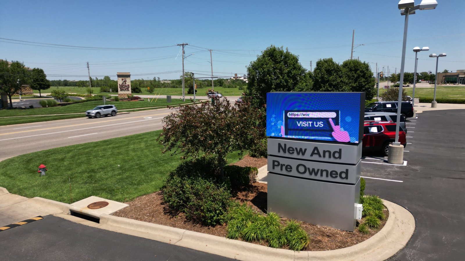 Car dealership roadside sign with “New And Pre Owned” and an LED electronic message center reading “Visit Us”; parked cars nearby.