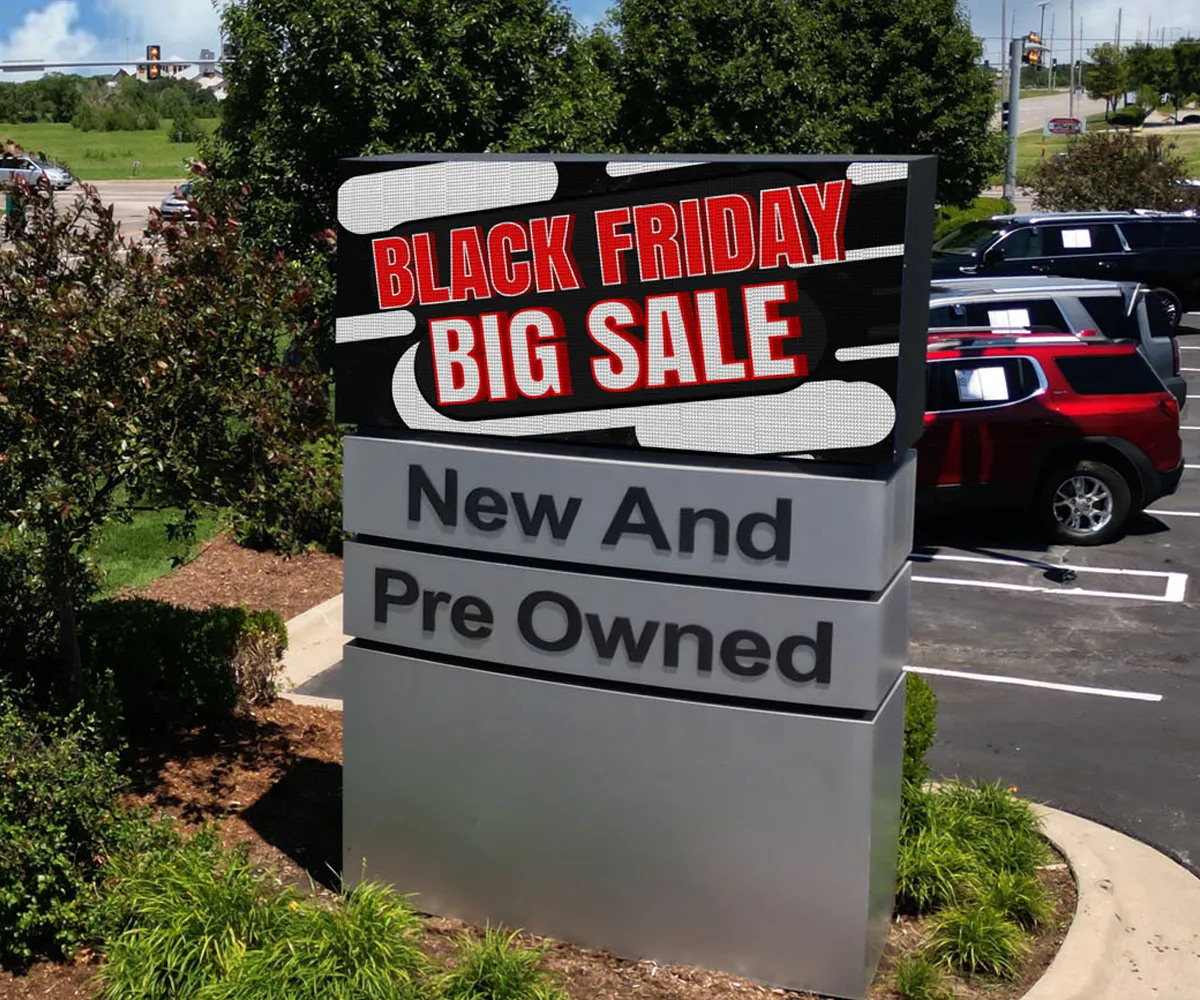 An outdoor high-brightness LED electronic message center displays "BLACK FRIDAY BIG SALE" above "New And Pre Owned" at a car dealership parking lot with multiple parked vehicles and surrounding greenery.