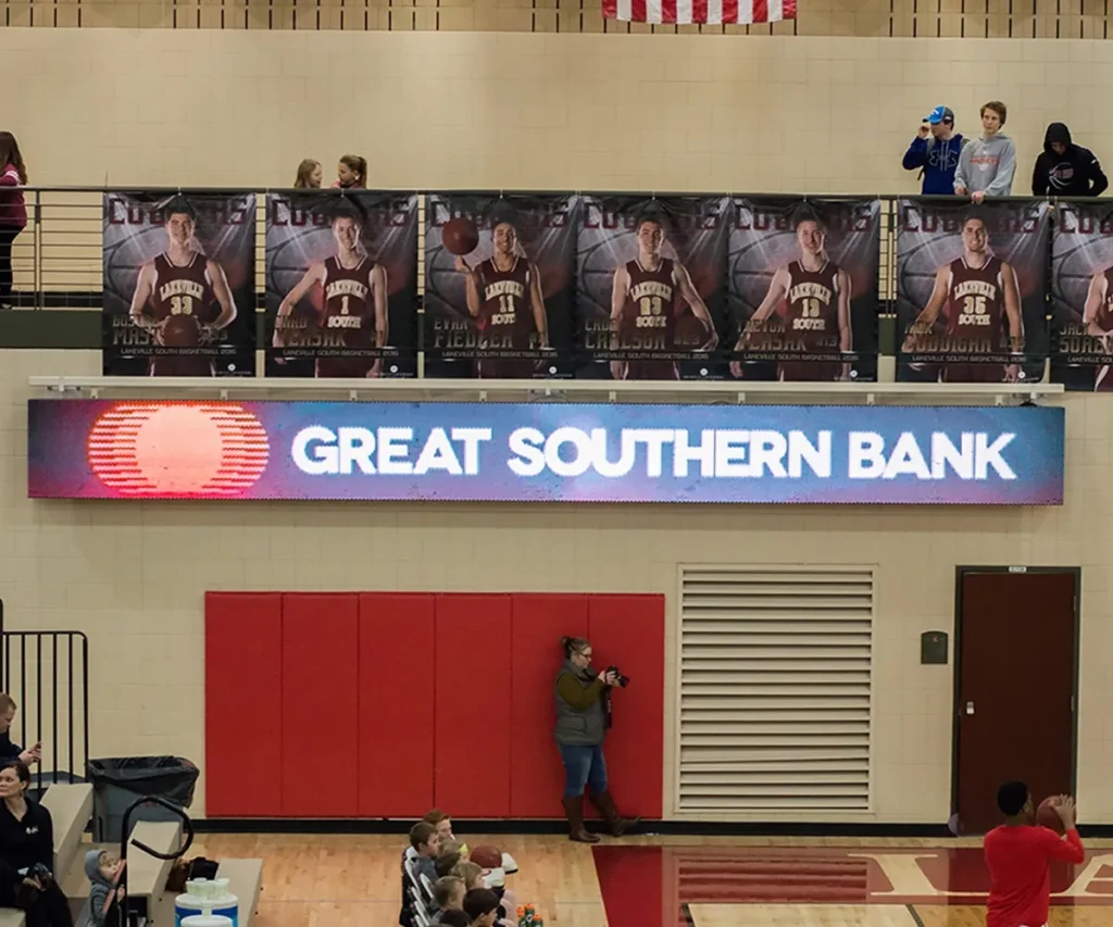 Large posters of basketball players are displayed above an LED electronic message center sign reading "Great Southern Bank" in a gymnasium. People are visible on the upper balcony, with a photographer standing below near the wall.