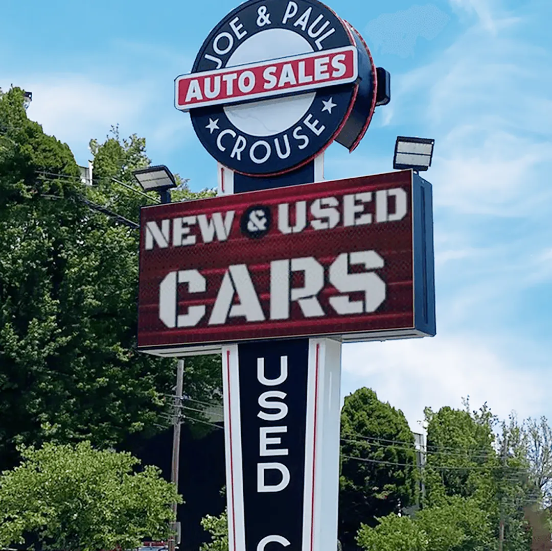 Tall aluminum commercial pylon sign for Joe & Paul Crouse Auto Sales, featuring an LED electronic message center displaying "NEW & USED CARS" in high-brightness lighting; trees and blue sky visible behind the signage.