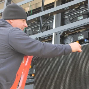 A person in a black hoodie and beanie stands on a ladder, installing or servicing an electronic message center with visible solid-state LED modules and exposed wiring.