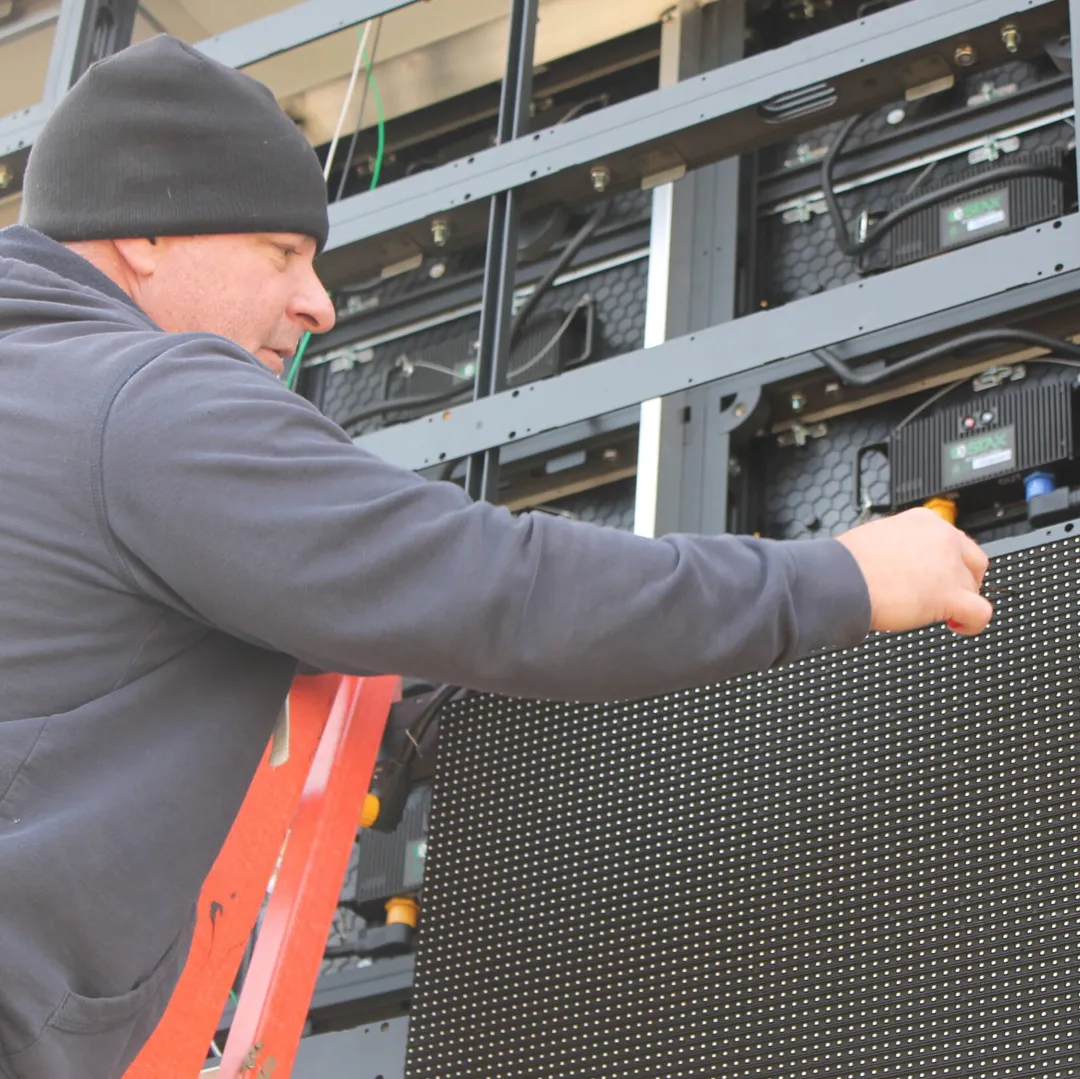 Technician on an orange ladder working on an open LED display at Kansas Lighting in Wichita, Kansas, with modules and internal components visible.
