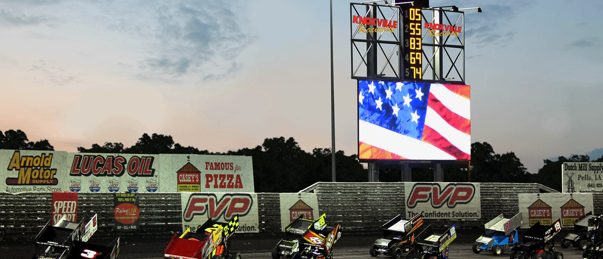 Several brightly colored race cars line up on a dirt track at dusk. Behind them, a high-brightness solid-state LED scoreboard and an electronic message center show the American flag. Aluminum-framed advertising banners are placed along the perimeter fence.