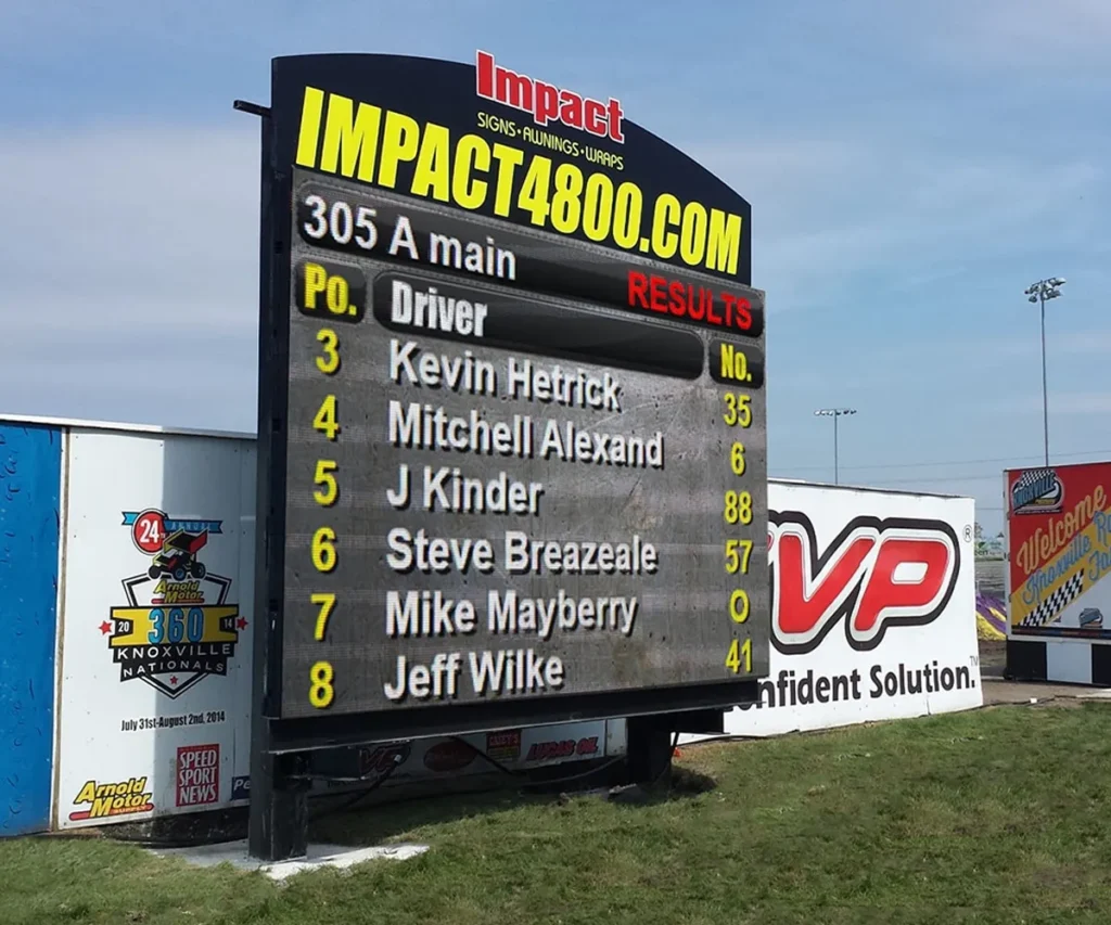 High-brightness outdoor LED electronic message center displays race results with driver positions, names, and numbers including Kevin Hetrick and Mitchell Alexan. Solid-state leaderboard features are housed in a weather-resistant aluminum frame. Commercial banners and advertisements appear in the background.