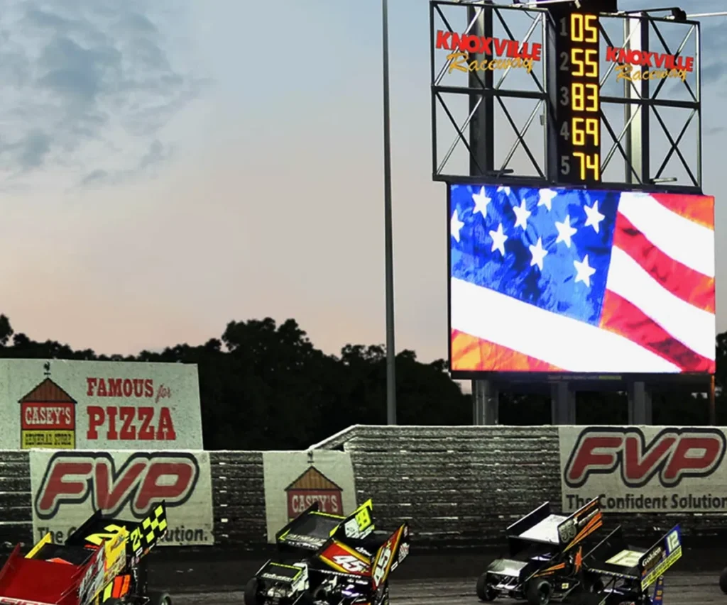 Dirt track racecars speed by grandstands at Knoxville Raceway, with a solid-state, high-brightness LED scoreboard and an aluminum electronic message center displaying the American flag in the background.