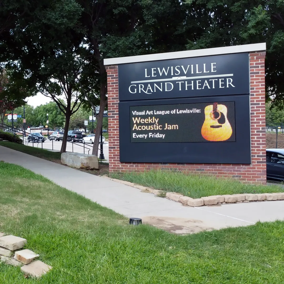 An LED electronic message center for Lewisville Grand Theater shows “Weekly Acoustic Jam Every Friday” alongside a guitar graphic. The solid-state display is set against a backdrop of trees, grass, and a concrete sidewalk.