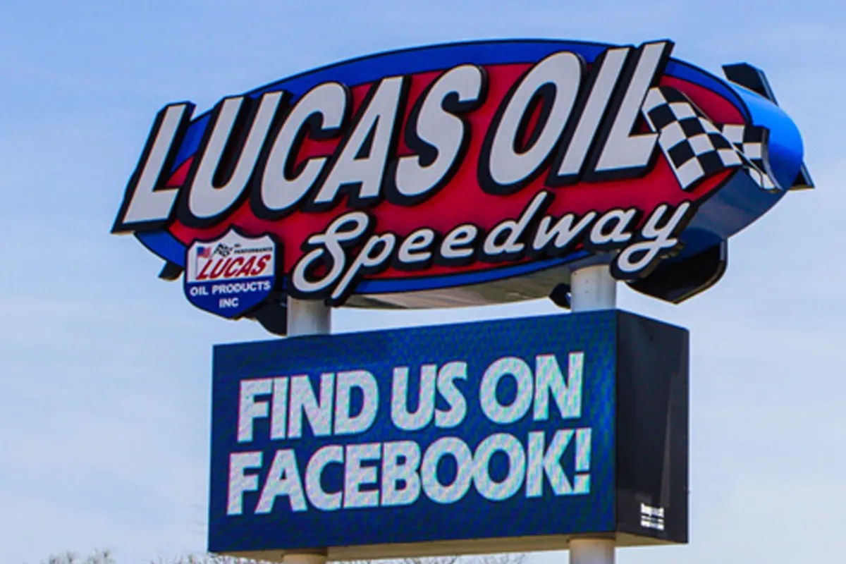 Lucas Oil Speedway commercial sign with checkered flag graphic; below, an outdoor high-brightness LED electronic message center displays “FIND US ON FACEBOOK!” Solid-state display and aluminum frame visible against blue sky backdrop.