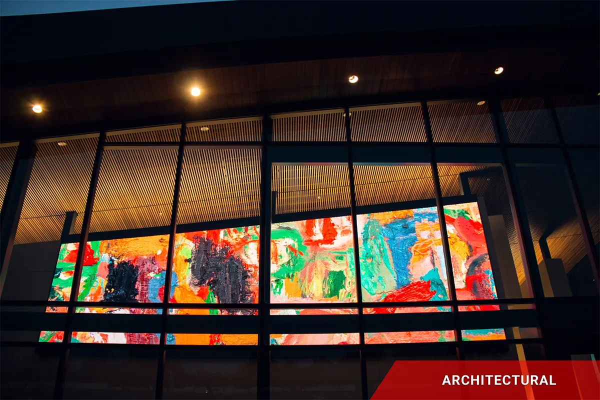 A vibrant, multi-colored abstract mural is visible through the windows of a modern building at night, illuminated by overhead warm lighting. In the bottom right corner, a high-brightness LED sign with solid-state technology displays the word “ARCHITECTURAL” in red.