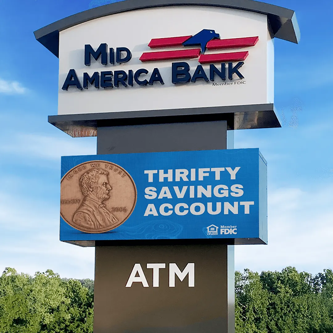 Aluminum commercial bank signage displays “MID AMERICA BANK” above an electronic message center advertising a “THRIFTY SAVINGS ACCOUNT” with penny graphic. A high-brightness LED panel at the bottom reads “ATM”. Trees and clear blue sky form the background.