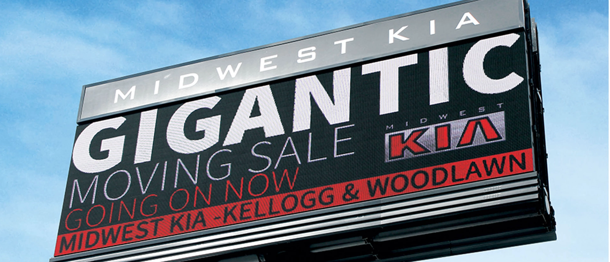 LED electronic message center displays "Gigantic Moving Sale" in bold, high-brightness lettering at Midwest Kia, with location details "Kellogg & Woodlawn." The phrase "Going on now" appears in orange text.