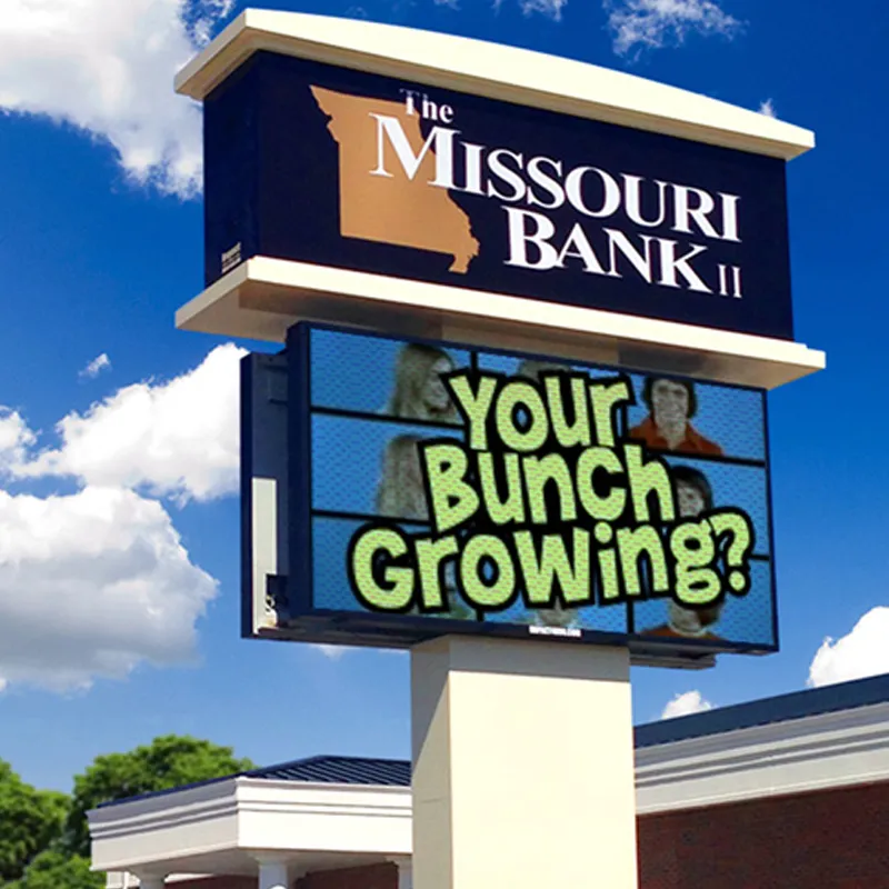 A Missouri Bank II commercial sign is mounted above an LED electronic message center displaying "Your Bunch Growing?" On the digital screen, photos of smiling people appear in the background, set against a clear blue sky.