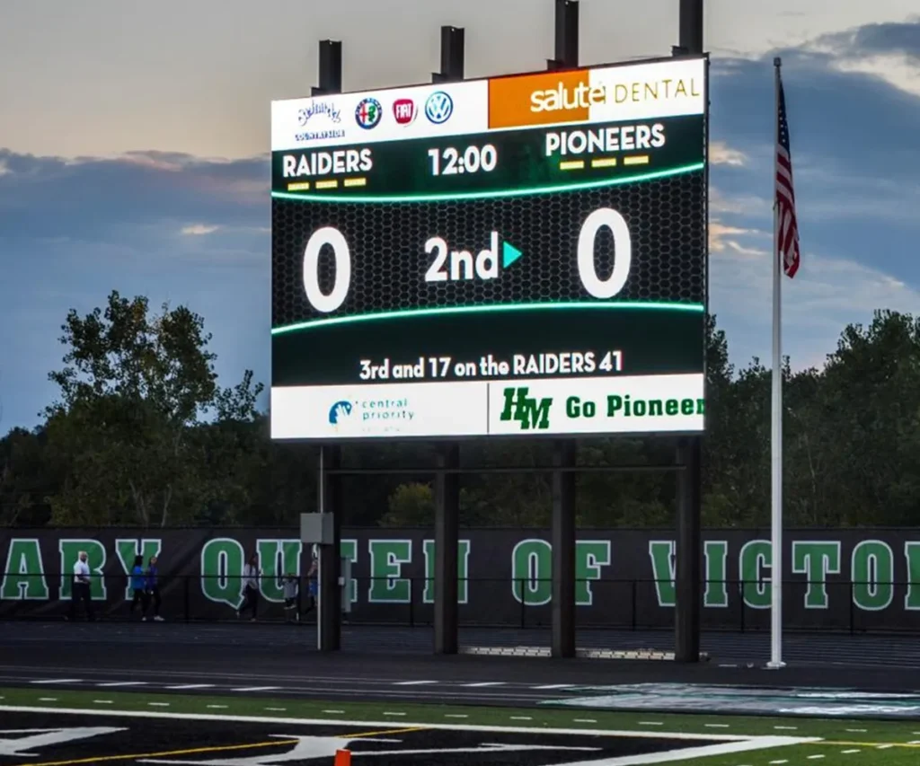 An LED electronic message center scoreboard displays Raiders 0, Pioneers 0, 2nd quarter with 12:00 on the clock. It indicates 3rd down and 17 at the Raiders’ 41-yard line. The solid-state sign overlooks an empty football field, with an American flag and trees in the background.