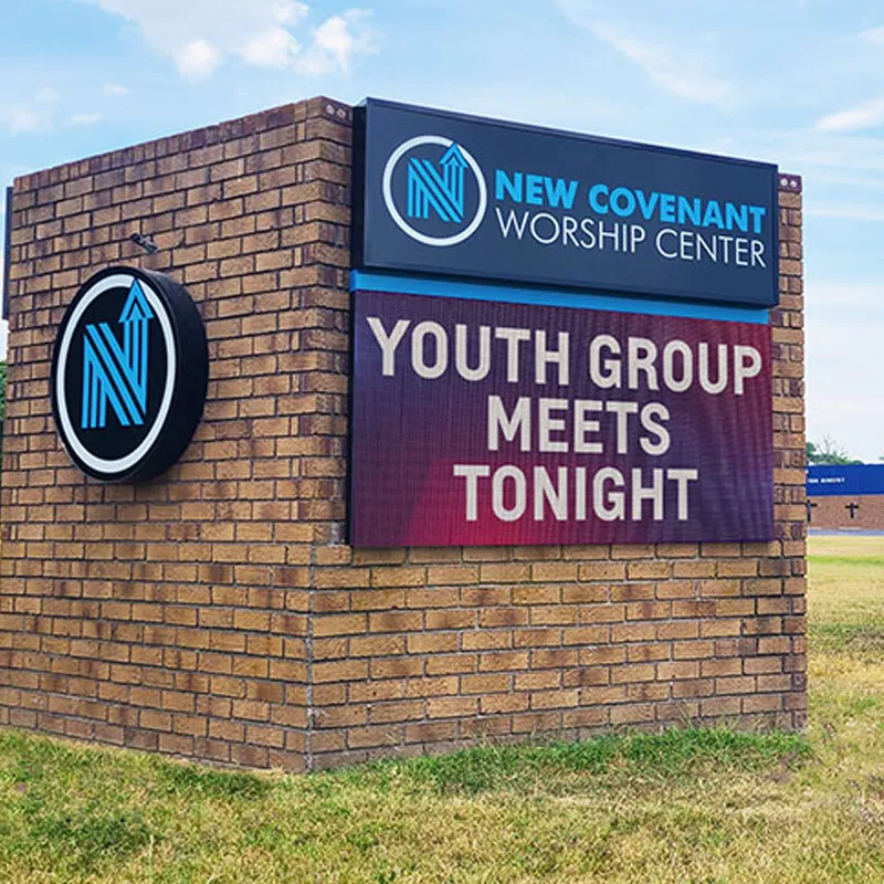 A brick monument sign features an LED electronic message center displaying "New Covenant Worship Center" and "Youth Group Meets Tonight," accompanied by a blue and white logo. The sign is set against a backdrop of grass and open sky.