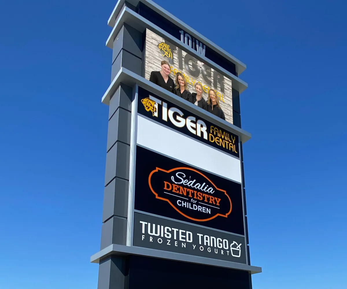 A tall aluminum signpost features an LED electronic message center displaying commercial signage for Tiger Family Dental, Sedalia Dentistry for Children, and Twisted Tango Frozen Yogurt beneath a clear blue sky. Near the top of the sign, a solid-state high-brightness display shows a photo of four smiling people.