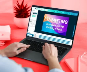 A person uses a laptop to design an LED sign slide displaying “PTA MEETING Thursday 7:30PM Parking in Back” in high-brightness text, simulating an electronic message center. A red plant and a notebook sit on the pink aluminum desk.